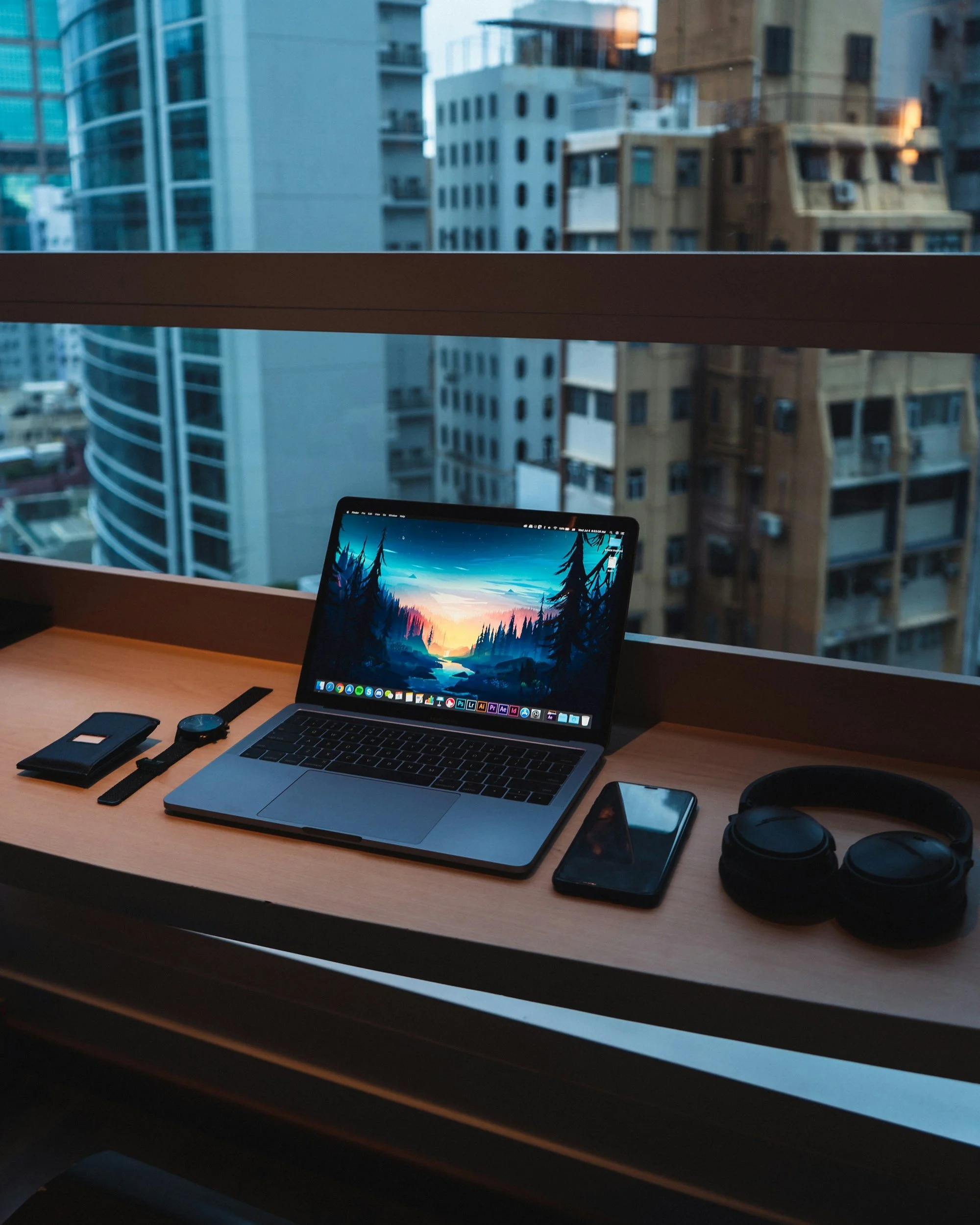 A workspace with a MacBook laptop, smartphone, headphones, watch, and wallet on a wooden desk, overlooking a cityscape from a high-rise window during dusk.