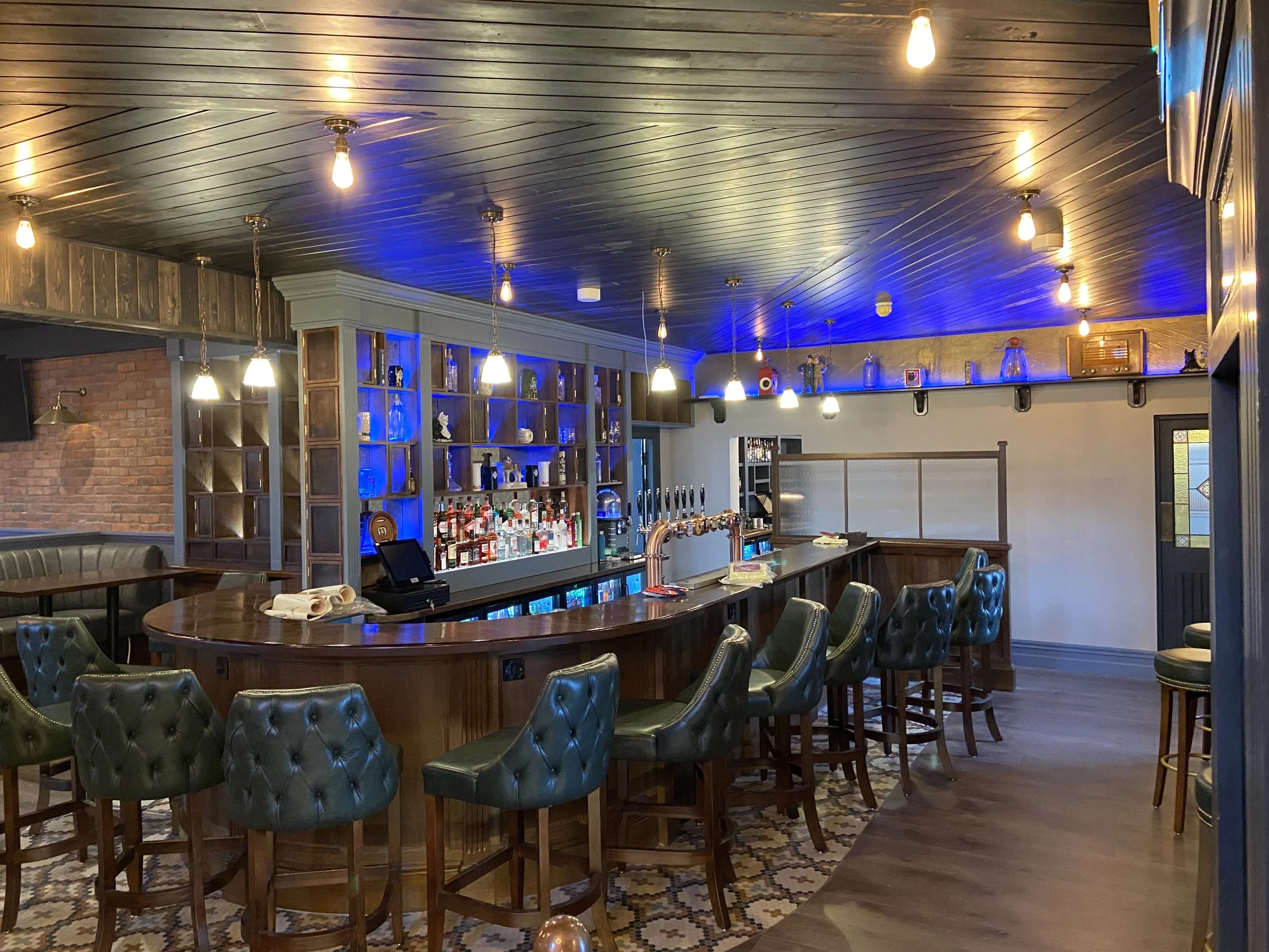 Empty bar with high green upholstered chairs around a wooden curved bar counter, liquor bottles and beer taps, with pendant lights hanging from a wooden ceiling; background has shelves with glassware and decorative items, and a wall with blue accent lighting.