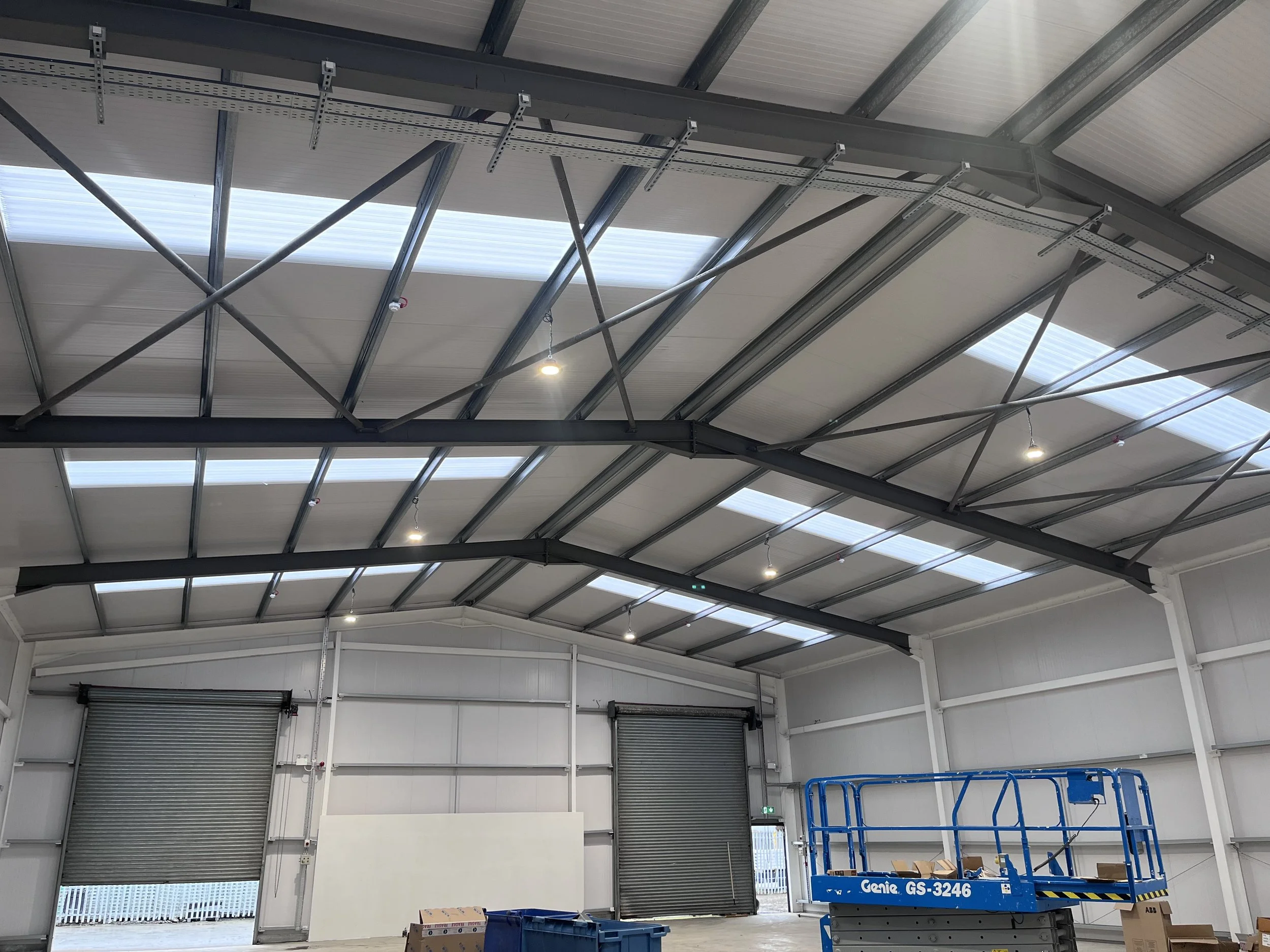 Interior of a metal warehouse with open garage doors, skylights in the ceiling, and a blue scissor lift.
