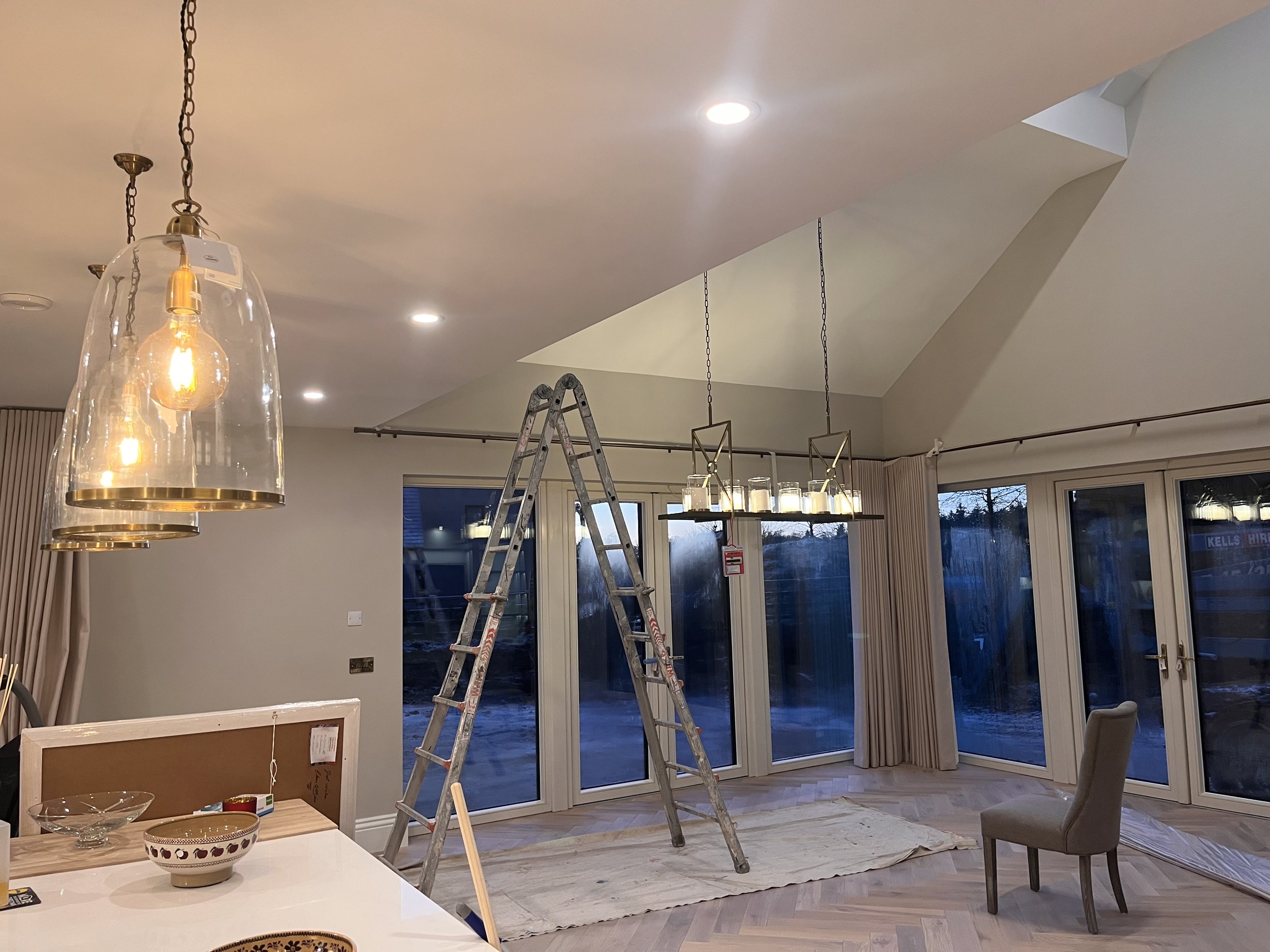 Living room with modern lighting fixtures, including a chandelier with candles, a hanging pendant light with two bulbs, a ladder, and a chair, during sunset or early evening outdoor view through large glass doors.