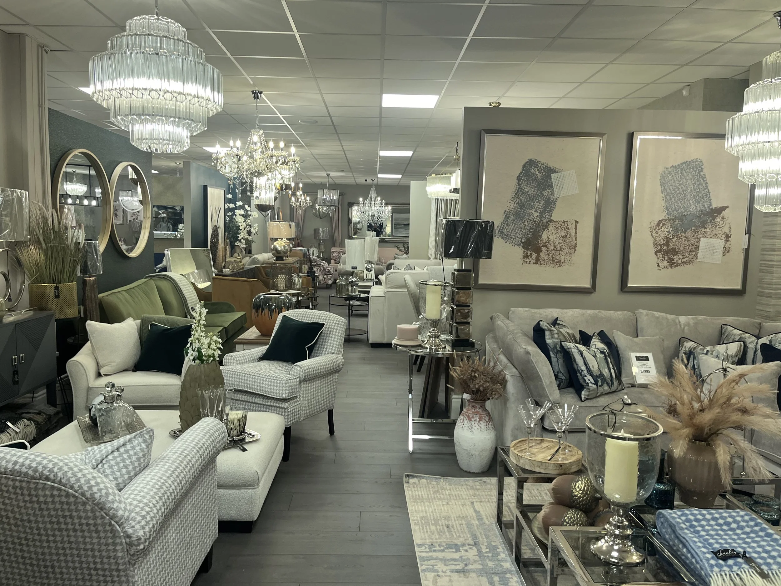 Interior of a furniture store with sofas, armchairs, tables, lamps, chandeliers, and decorative accents in neutral and muted colors.