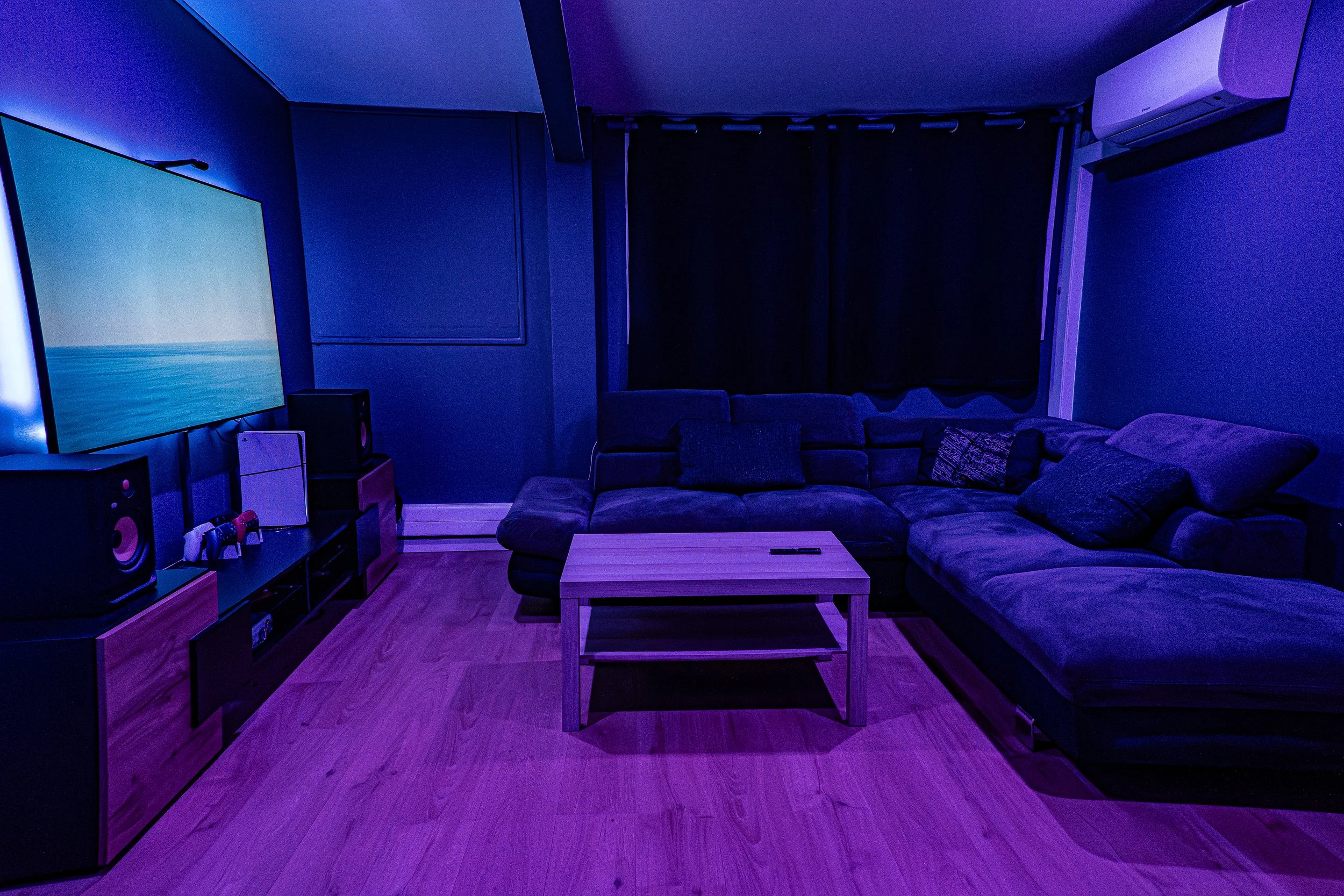 Salon avec canapé en coins, table en bois, télévision écran plat, haut-parleurs, appareils électroniques, voilages noirs, climatiseur, éclairage tamisé