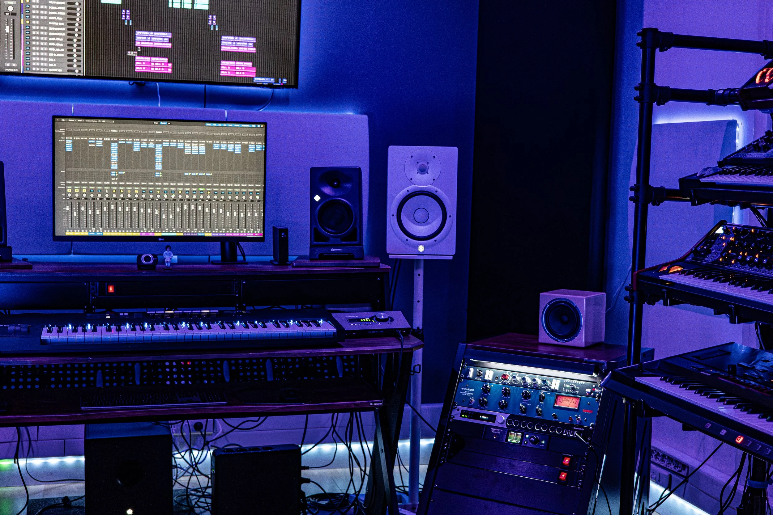 studio de musique NPL Studio avec plusieurs écrans, synthétiseurs, haut-parleurs, et équipements audio de haute qualité sous un éclairage bleu. 