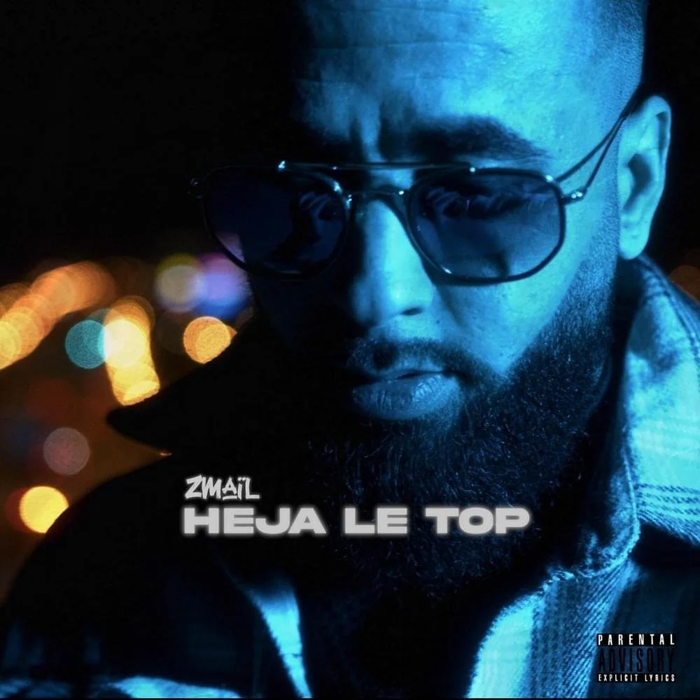 Portrait d'un homme portant des lunettes de soleil dans un environnement nocturne avec des lumières floues en arrière-plan, la légende 'ZMAIL HEJJA LE TOP' en bas de l'image.