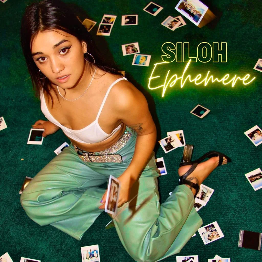 Une jeune femme assise sur un tapis vert, entourée de nombreuses photos de polaroid éparpillées autour d'elle. Elle porte un haut blanc, un pantalon vert pâle avec une ceinture à paillettes, et des chaussures à talons noirs. Il y a un signe lumineux 