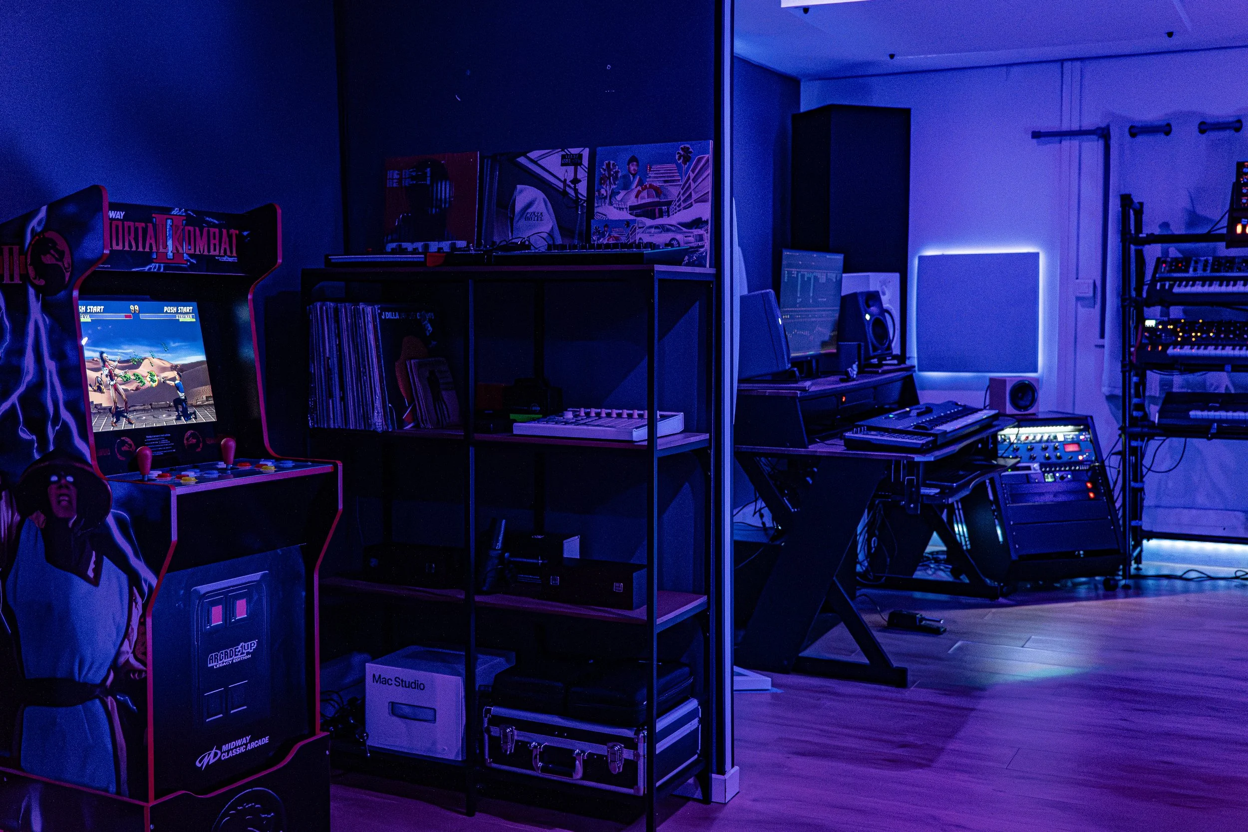 Une pièce sombre avec un coin de jeux vidéo, un ordinateur et un clavier, de nombreux équipements de musique, et une machine d'arcade "Mortal Kombat"