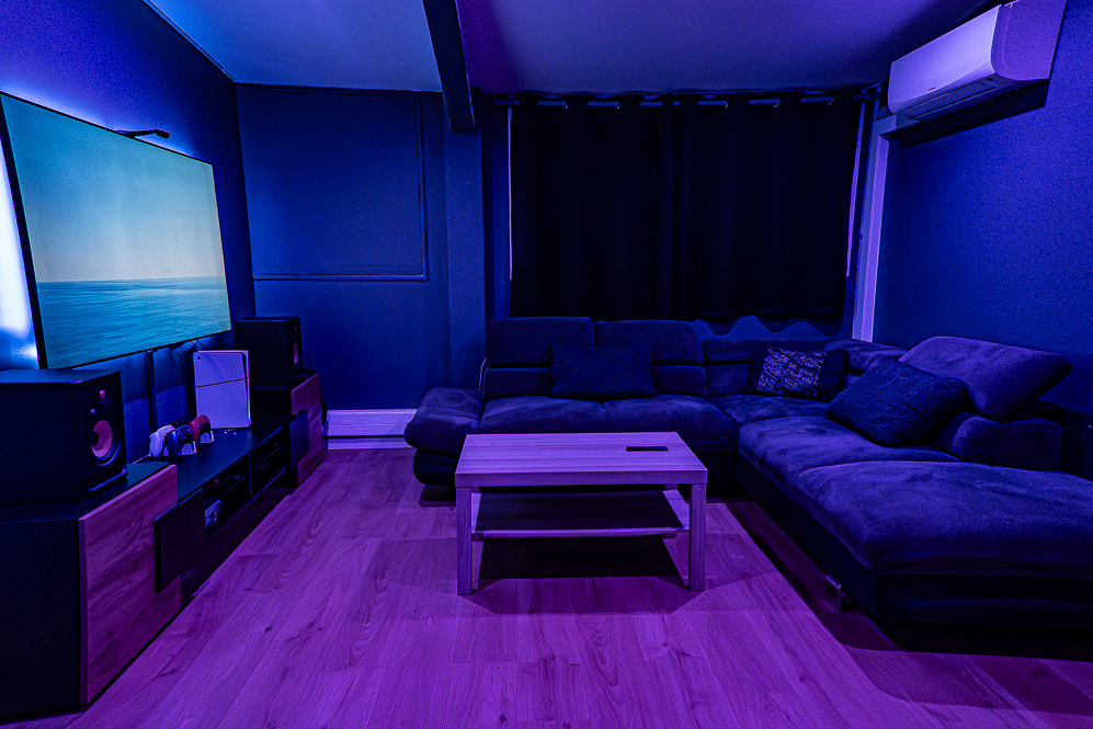 Salon sombre avec un canapé en U, table basse en bois, mur avec écran de télévision, et climatiseur mural, éclairé par une lumière bleue.