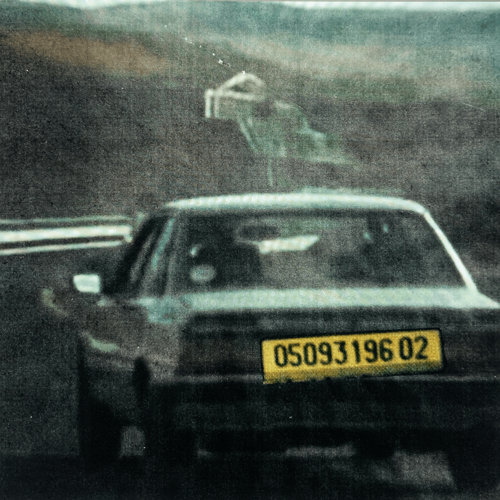 Une voiture de police noire vue de l'arrière, avec une plaque d'immatriculation jaune et noire. Une arme pointée au-delà du toit de la voiture, semblant être une arme à feu dans la main d'une personne. L'image est floue et a un aspect granuleux.