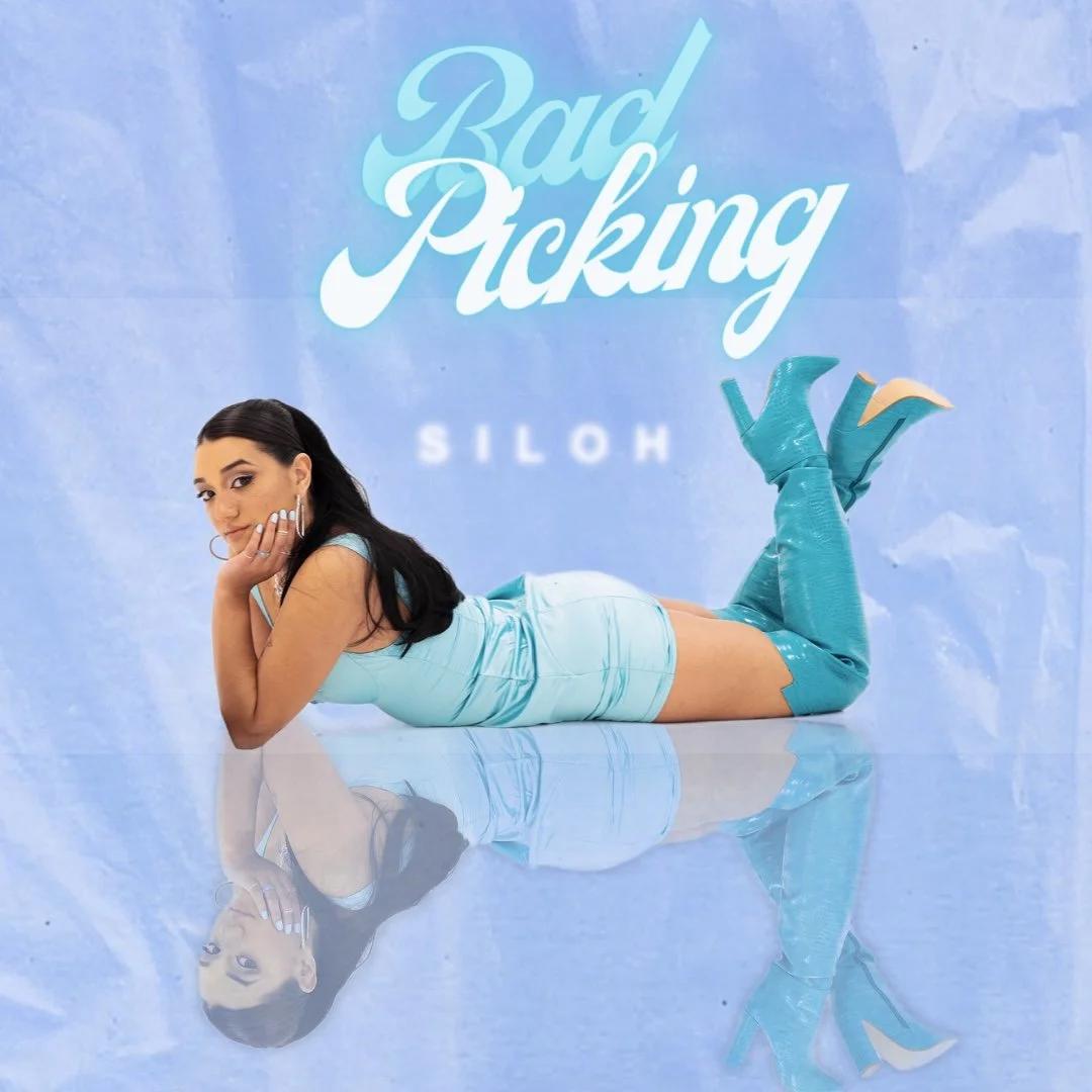 Une femme allongée sur le ventre, portant une robe et des bottes hautes en cuir bleu, regarde vers l'avant. Le fond montre le titre 'Bad Picking' en grandes lettres blanches sur un fond bleu. La réflexion de la femme est visible sur le sol brillant.