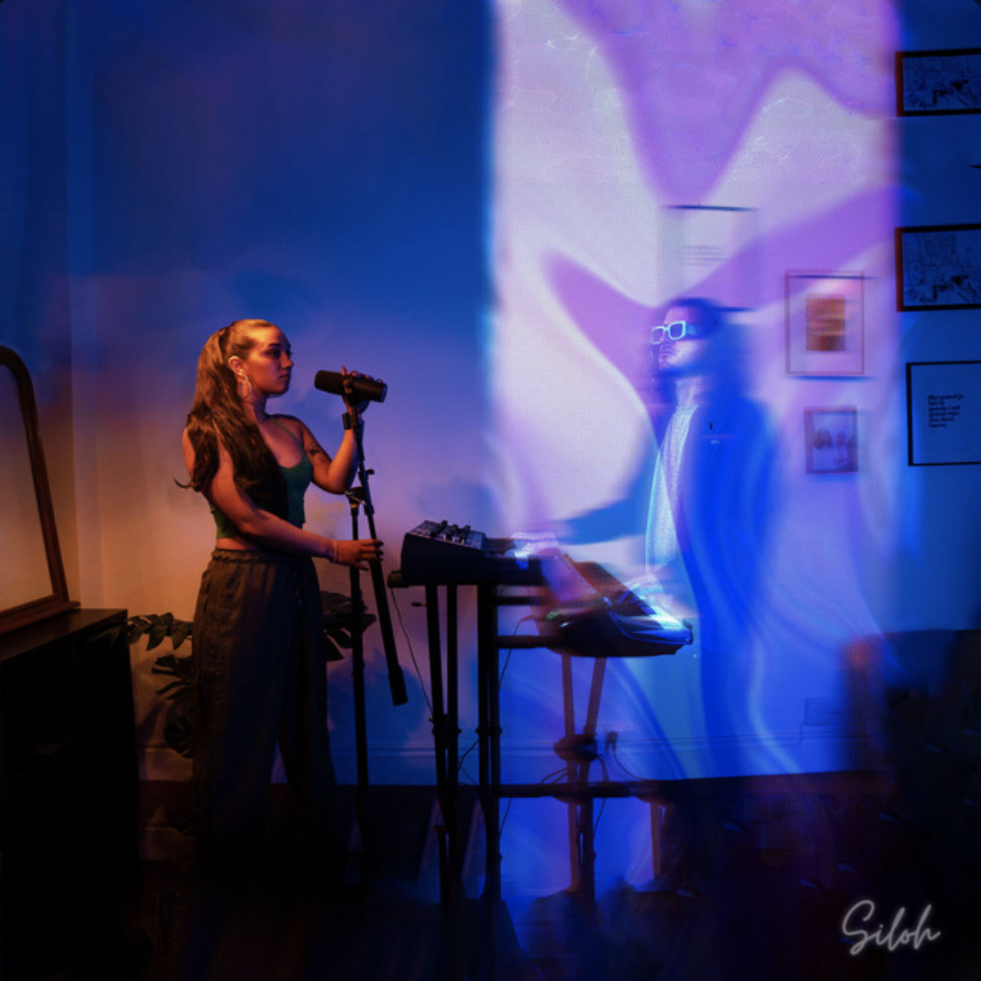 Deux jeunes femmes dans une pièce, une chantant à un microphone, l'autre derrière un clavier, avec des lumières colorées et un effet de mouvement sur la scène.