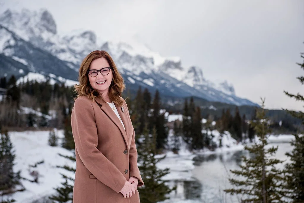 A smiling woman with glasses and shoulder-length wavy hair, wearing a tan coat, standing outdoors in a snowy mountain landscape with trees and a river in the background.