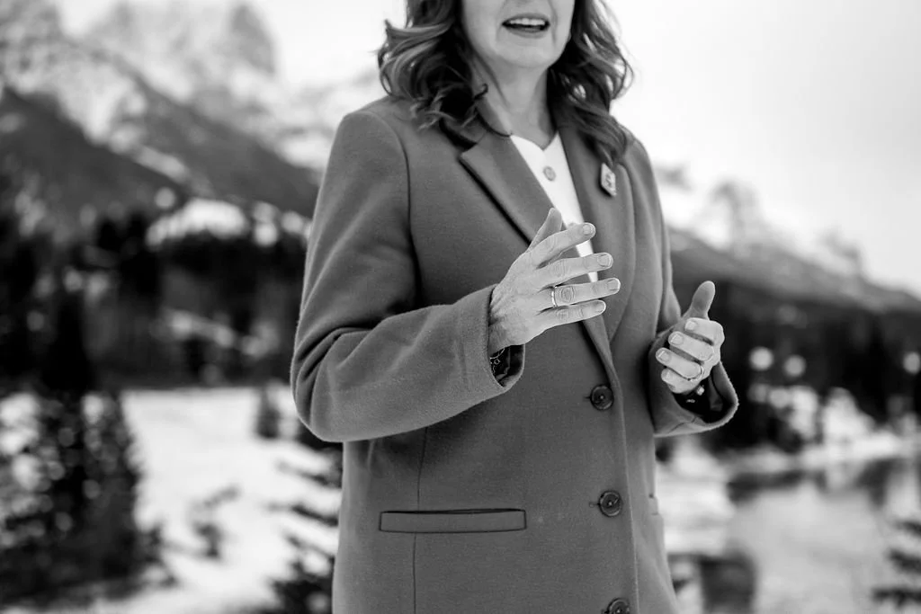 A woman in a coat speaking outdoors with a snowy mountainous landscape in the background, black and white photo.