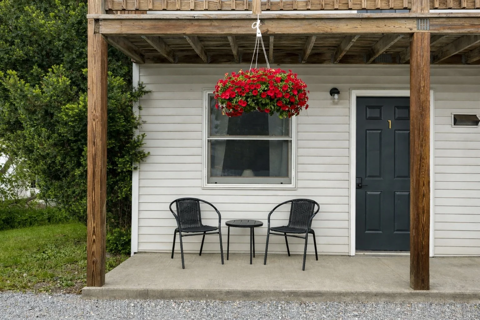 A white house exterior with a black front door numbered 1, a window, two black chairs with a small round table between them on a concrete porch, a hanging red flower basket, and a wooden deck above.