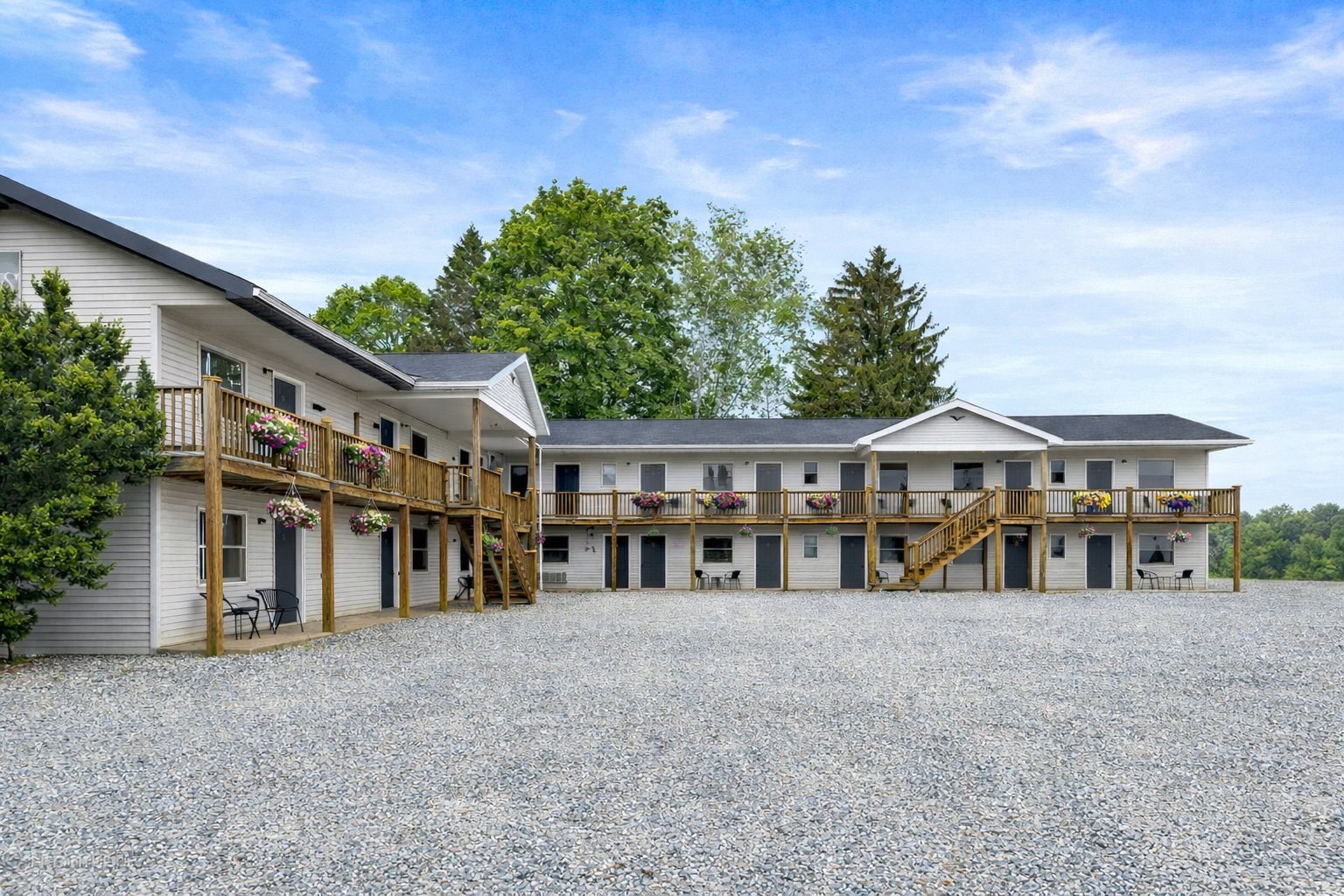 Two-story apartment building with white siding, black doors, and a large wooden deck with flower boxes. There are stairs leading to the upper deck and small outdoor seating areas below. Gravel parking lot in front and green trees in the background un
