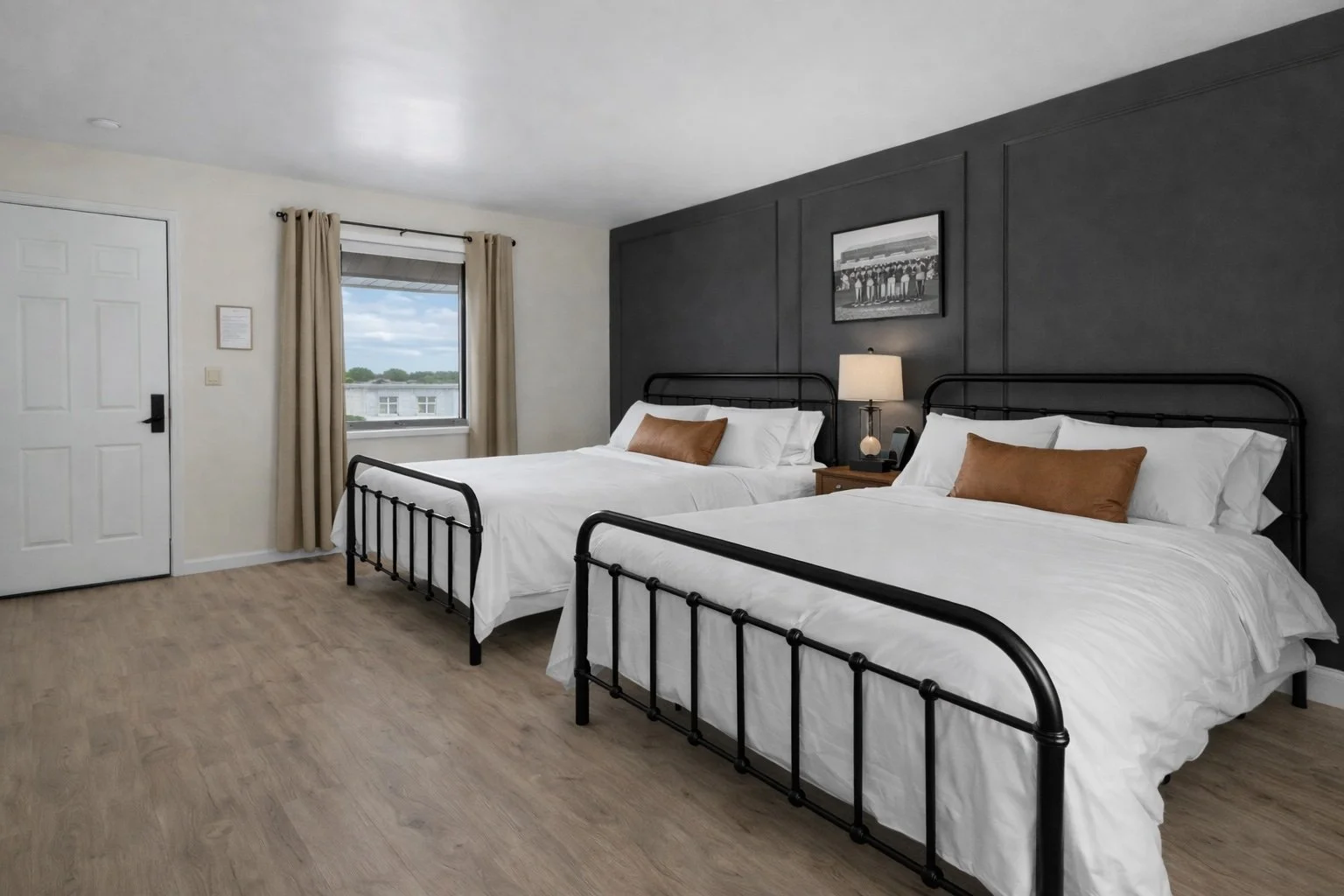 A hotel room with two twin beds with black metal frames, each with white bedding and brown pillows. A nightstand with a lamp and a phone is between the beds, against a gray accent wall. There is a window with beige curtains showing a view of a cloudy
