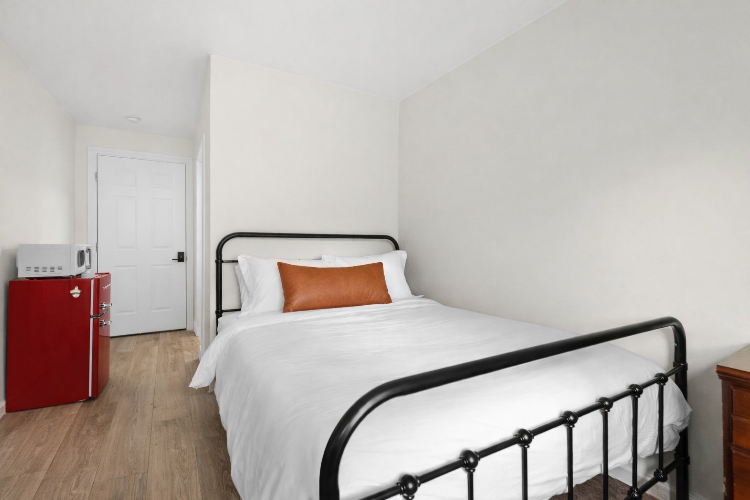 Comfortable bed with white linens and a rust-colored pillow in a minimalist bedroom with light-colored walls, wooden floor, and a small red refrigerator with a microwave on top near the door.