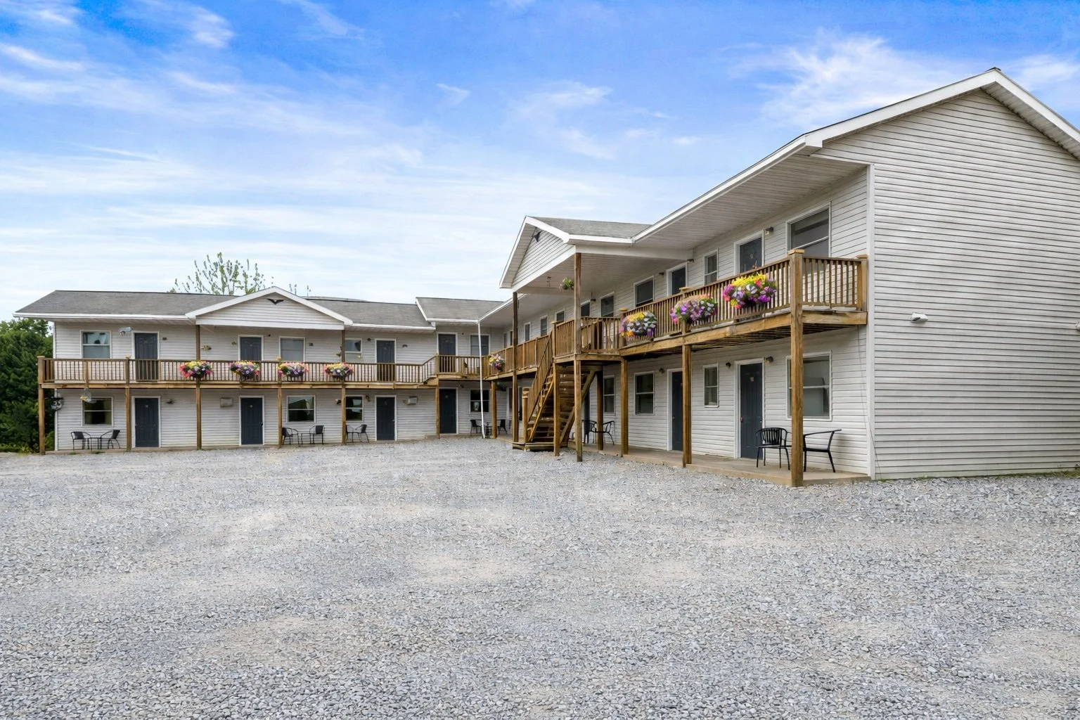 Exterior view of a two-story white apartment building with a gravel parking area, featuring balconies decorated with flower boxes, and outdoor seating.