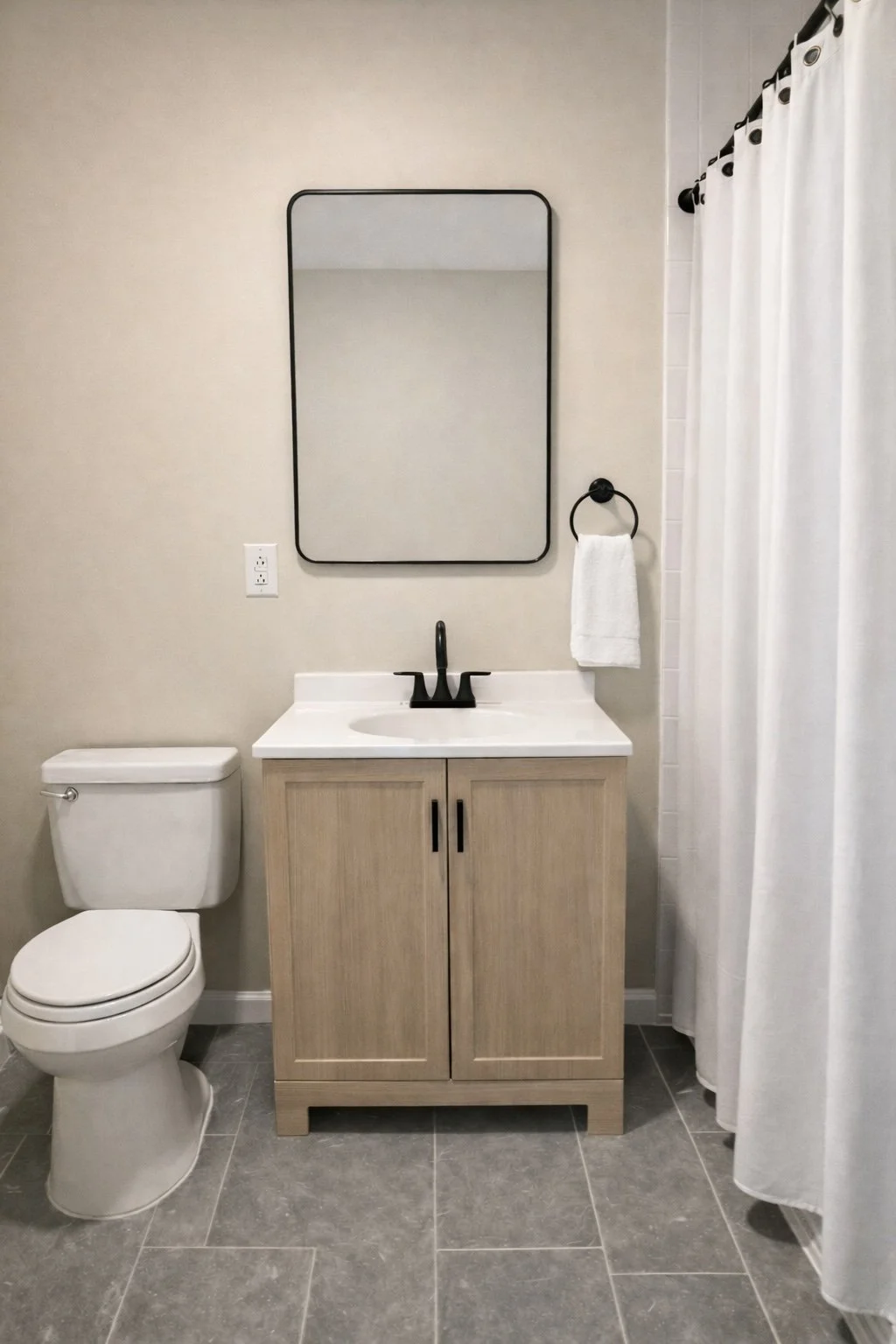 Bathroom with toilet, wooden vanity with black faucet, oval mirror, white towel on black ring, and white shower curtain.
