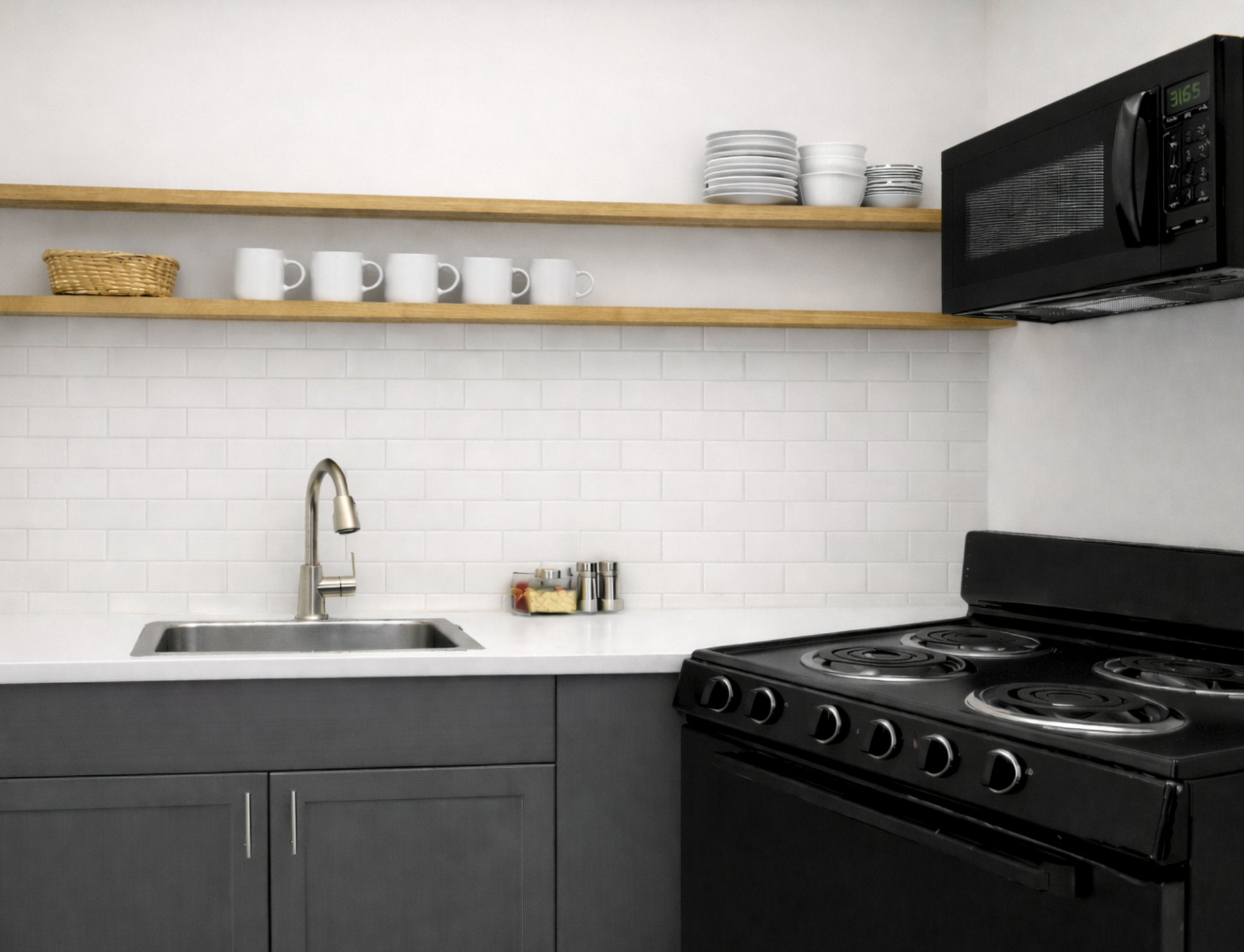 Modern kitchen with white subway tile backsplash, black stove, microwave, gray cabinets, a stainless steel sink, open wooden shelves with white dishes, dark countertops, and a small container with spices.