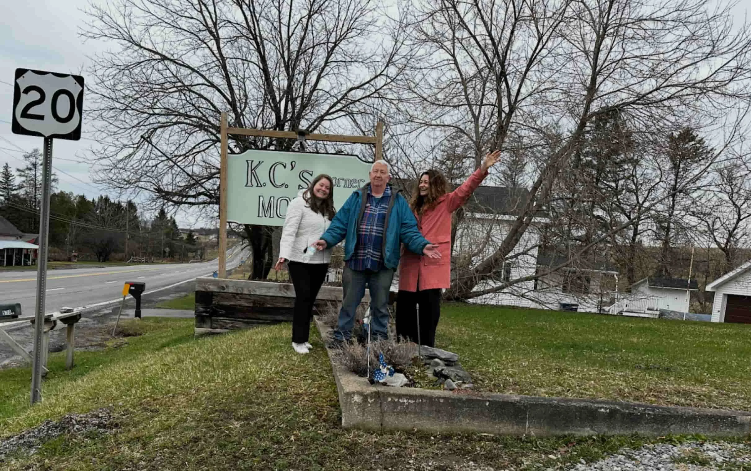 Three people standing on a grassy area near a road, smiling and posing for the camera with trees and houses in the background.