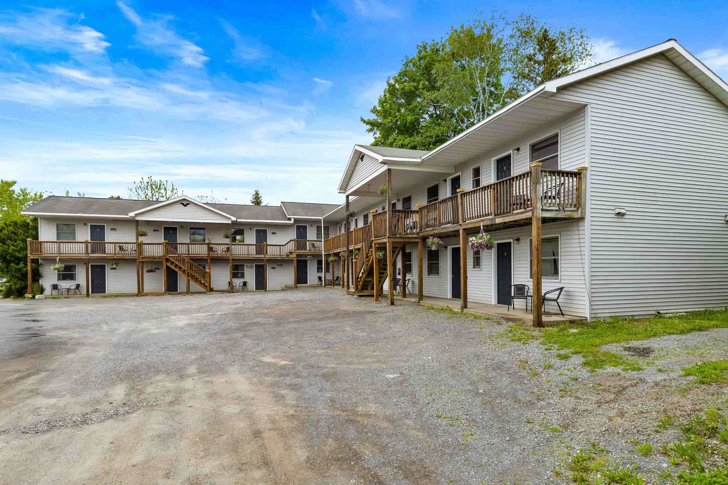 Two-story apartment buildings with exterior staircases, balconies, and a gravel parking area under a blue sky with some clouds.