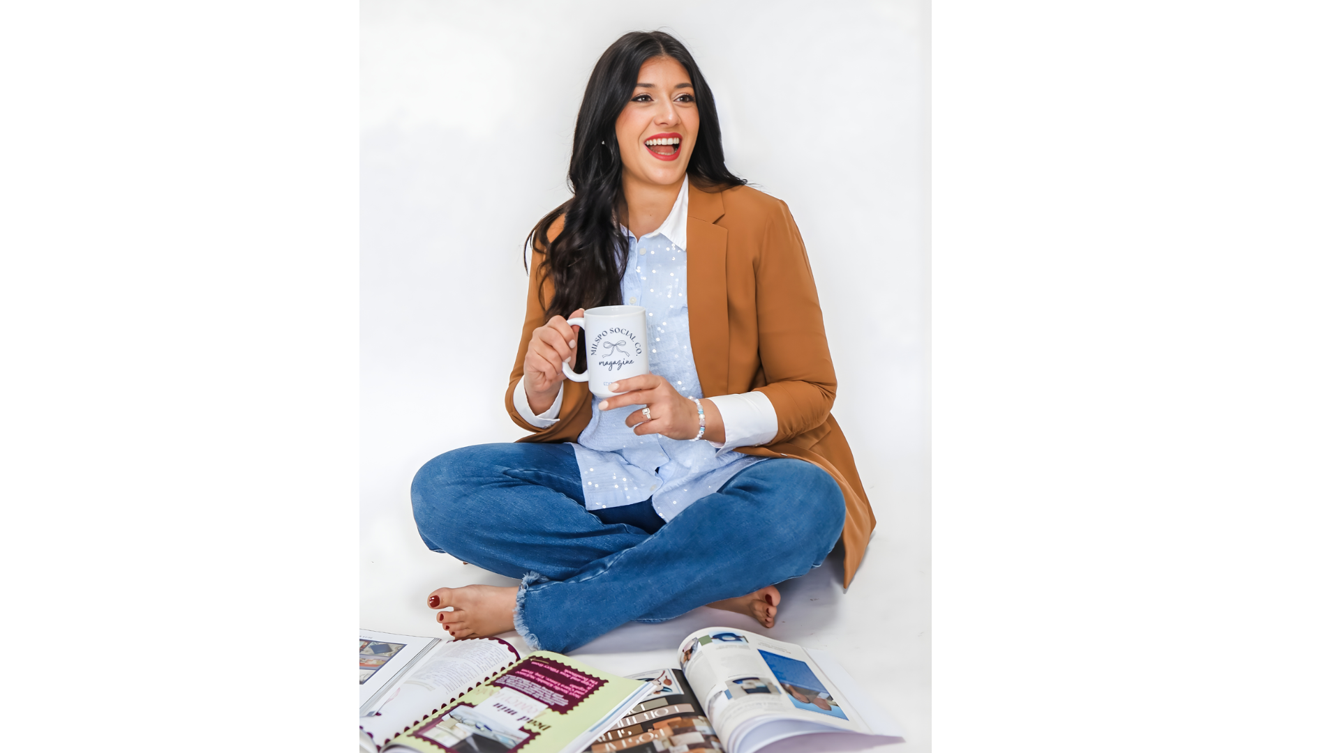 A woman sitting cross-legged on the floor, holding a white mug, smiling, surrounded by magazines and books, against a plain white background.