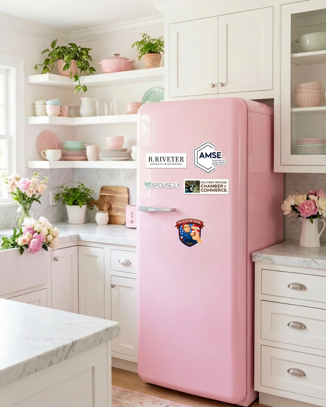 Pink retro-style refrigerator with various stickers on the front in a bright white kitchen with open shelves holding pink, white, and mint green dishes, marble countertops, and flowers in vases.