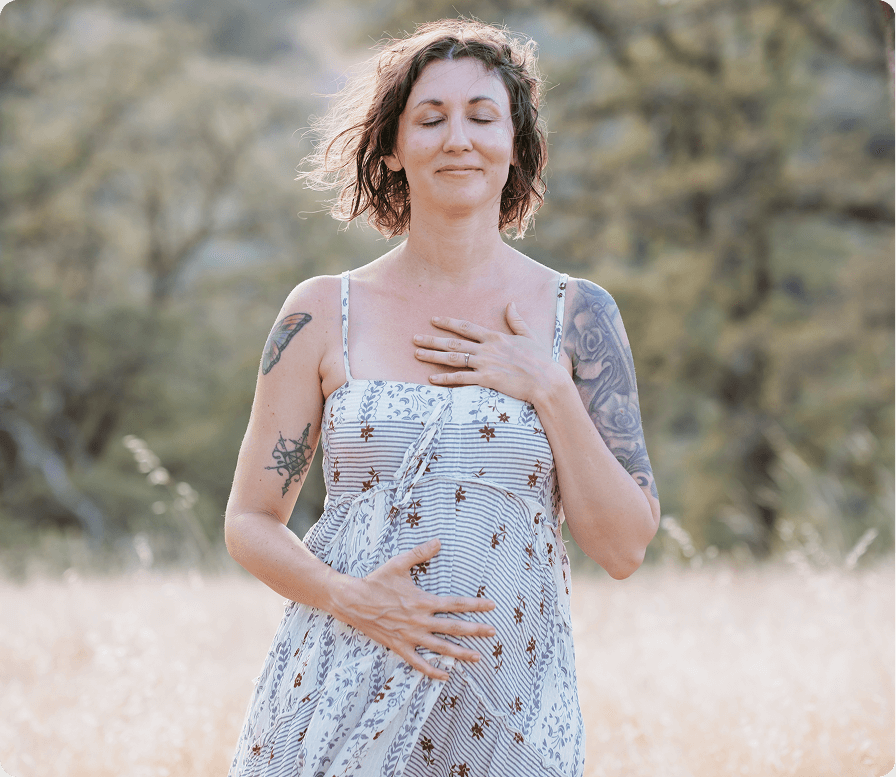 Elena Wolfbrandt-Michels, wearing a sleeveless dress with a floral pattern, standing outdoors in a field with trees in the background. She has her eyes closed and one hand on her chest and the other on her belly.