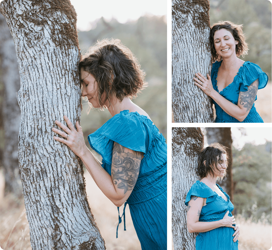 Elena in a blue dress, standing outdoors hugging a tree, smiling, and holding her belly as a somatic practice.