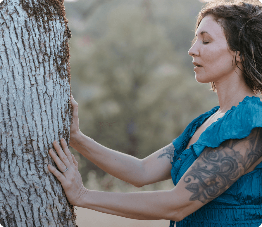 Elena Wolfbrandt-Michels, with dark hair and tattoos on her arm gently touches a tree trunk with her hands
