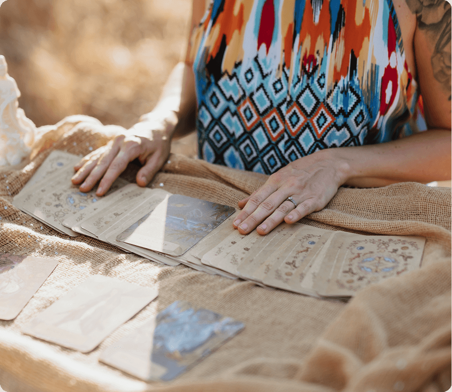 Person wearing a colorful patterned shirt flipping through tarot cards on a burlap cloth outdoors.