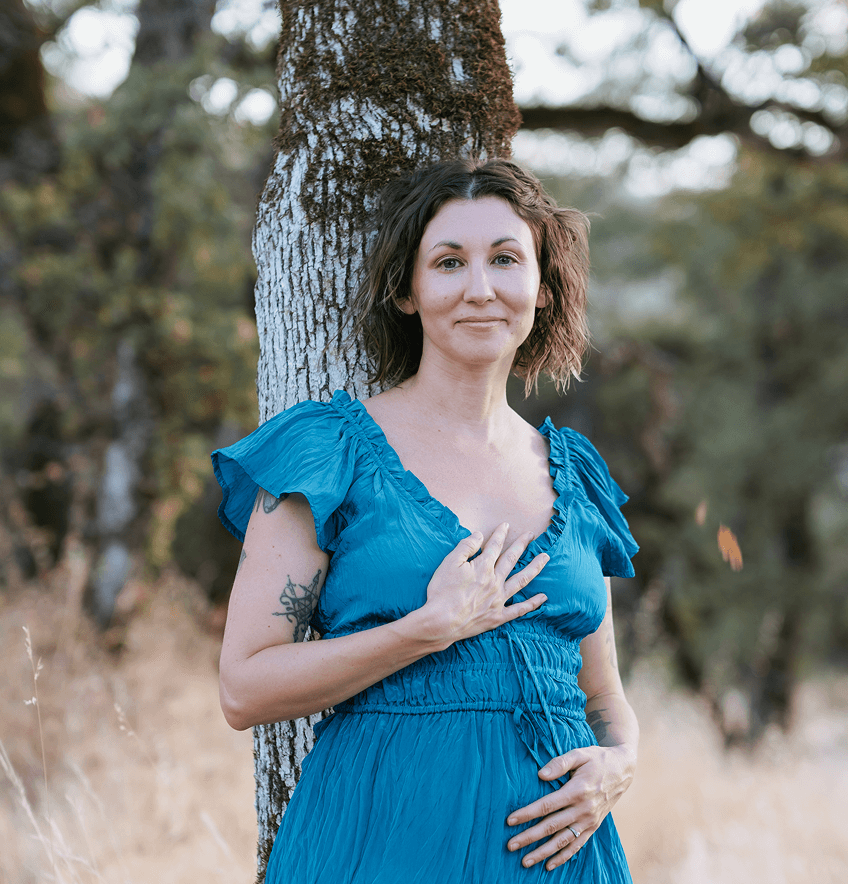 Elena, owner of Earth Body Oracle, wearing a blue dress, standing outdoors and leaning against a tree with her hand on her chest and stomach, taking a deep breath and feeling into her body.