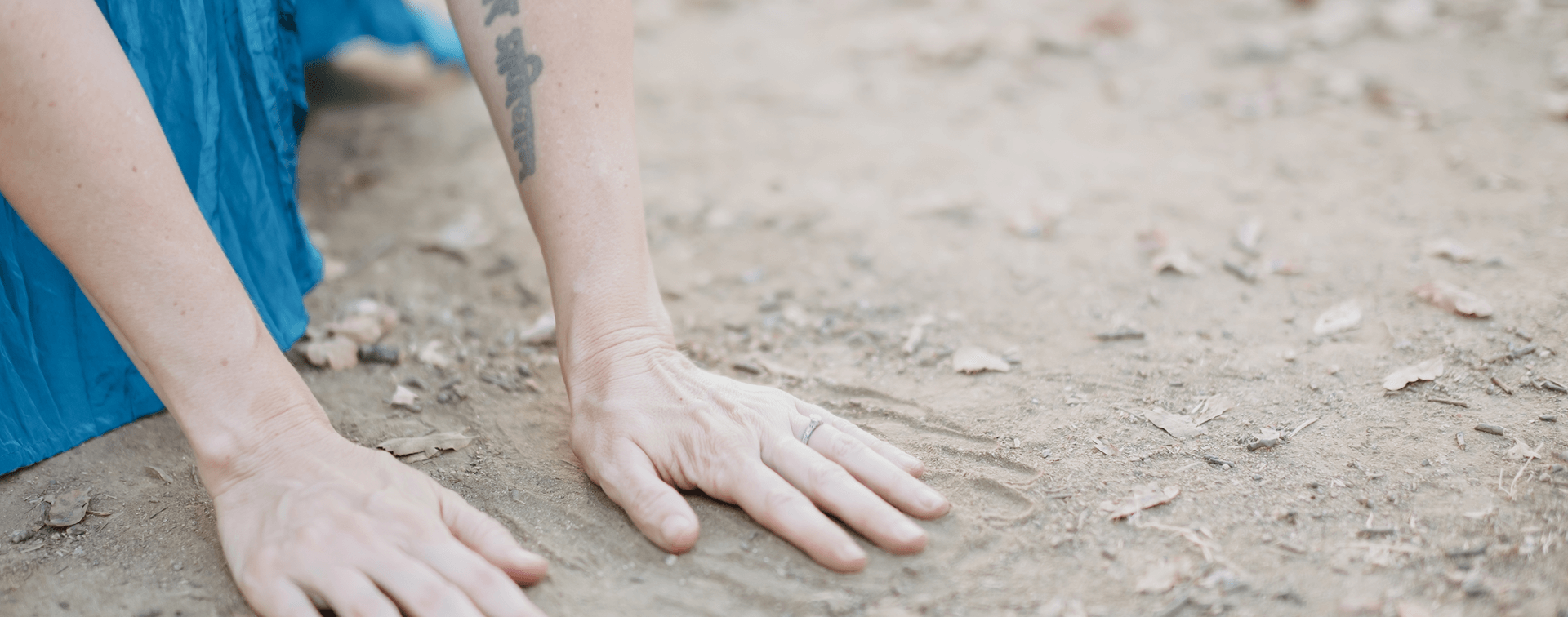 Elena, with tattoo on arm and wedding ring, has her hands in the dirt as she does an ecstatic dance.