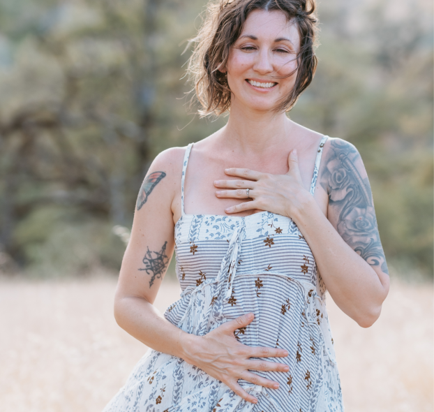 A smiling woman (Elena Wolfbrandt-Michels) in a blue and white sundress with tattoos on her arms, standing outdoors in a natural setting, doing a qi gong practice.