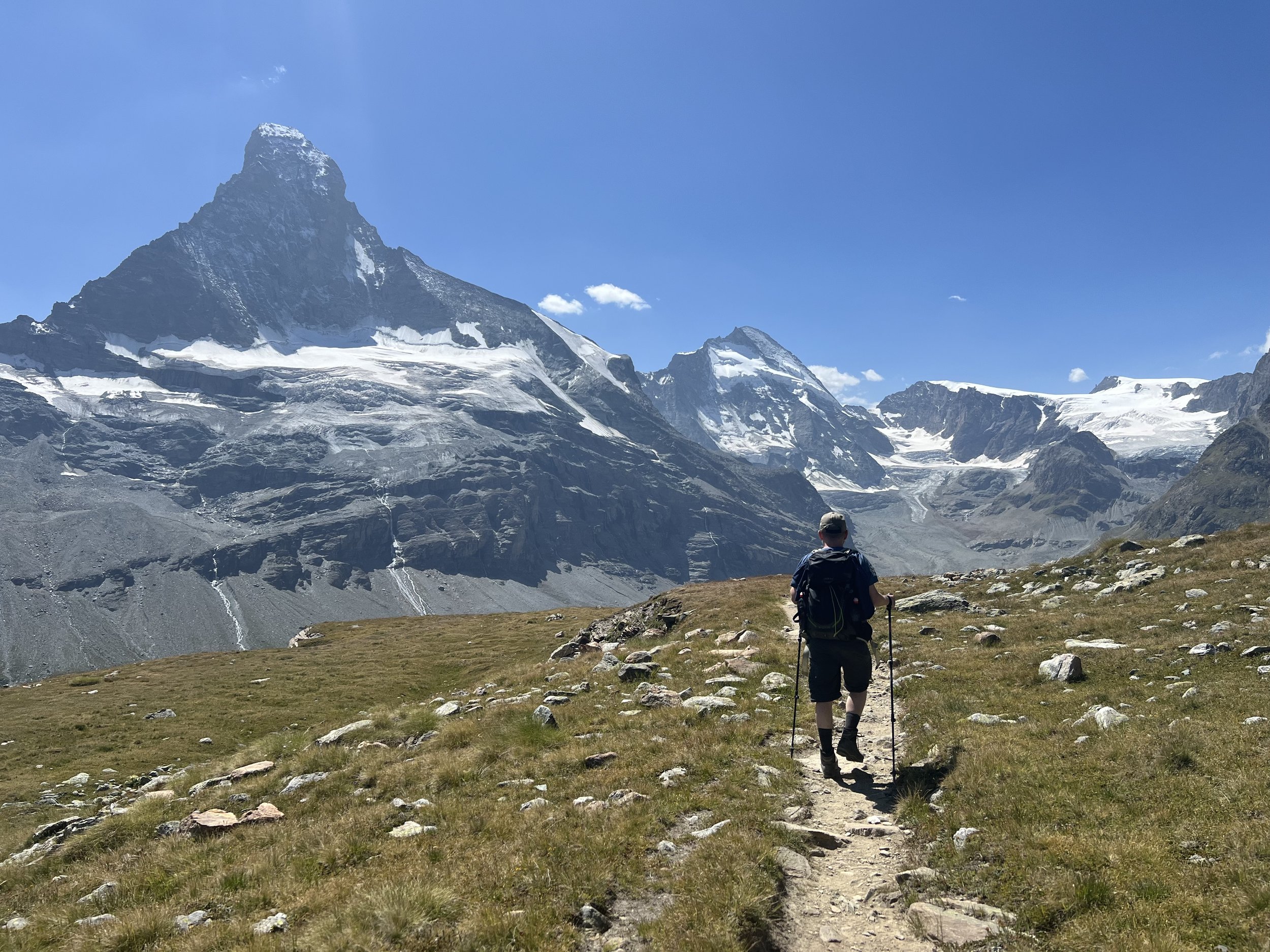 Ein Wanderer mit Rucksack und Wanderstöcken wandert auf einem Pfad durch eine bergige Landschaft mit schneebedeckten Gipfeln im Hintergrund unter einem blauen Himmel.