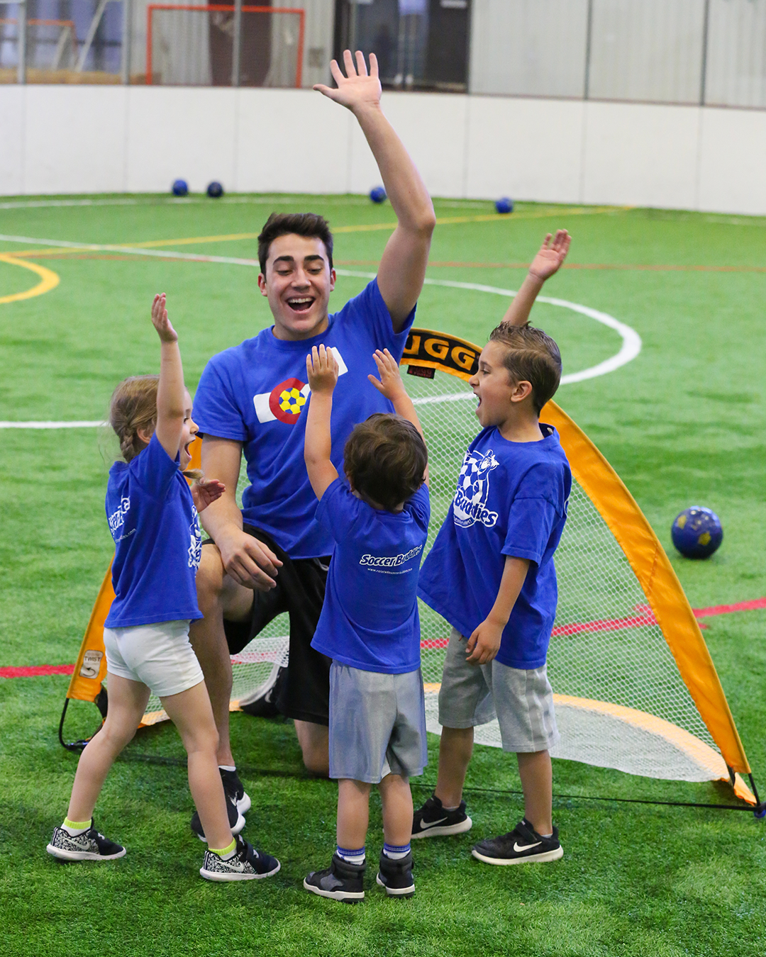 A young male coach with short dark hair smiling and raising his arm, surrounded by four young children on a green indoor soccer field, celebrating near a small orange soccer goal, with several small soccer balls in the background.