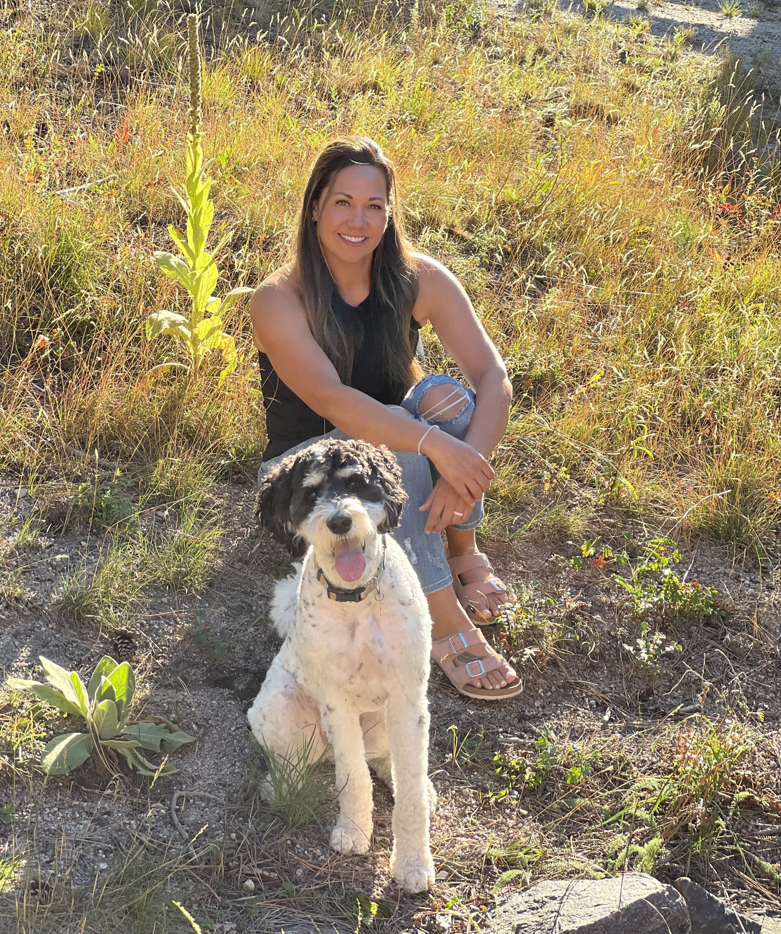 A woman with long brown hair wearing a black sleeveless top and ripped jeans is sitting on a hillside with her leg bent, smiling at the camera. She is beside a black and white dog with curly fur, sitting on the ground, looking happy with its tongue out. The background is a grassy field with some plants and sunlight.