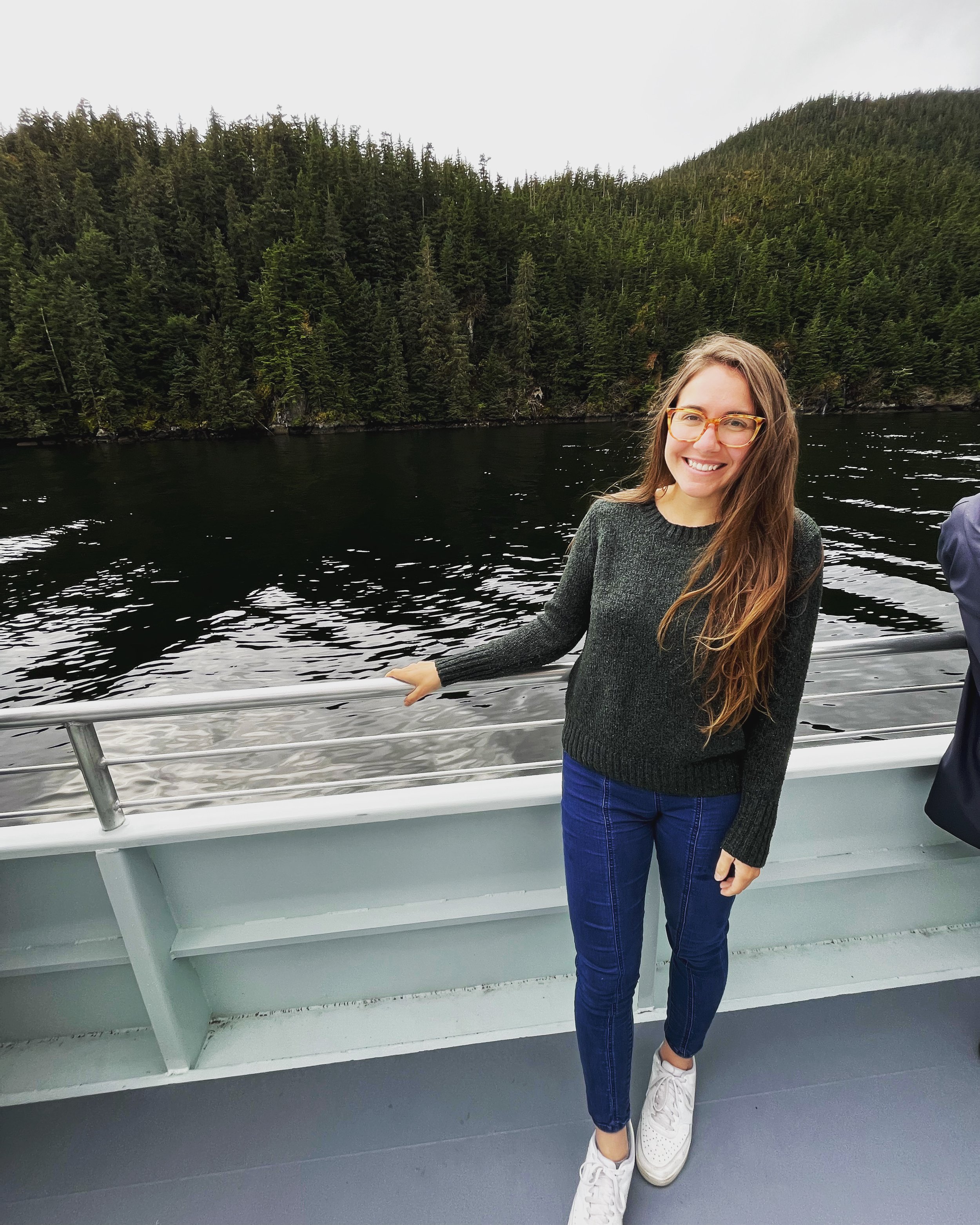 A woman with long brown hair, wearing glasses, a dark sweater, and blue pants, smiling on a boat with a forested mountain and dark water in the background.
