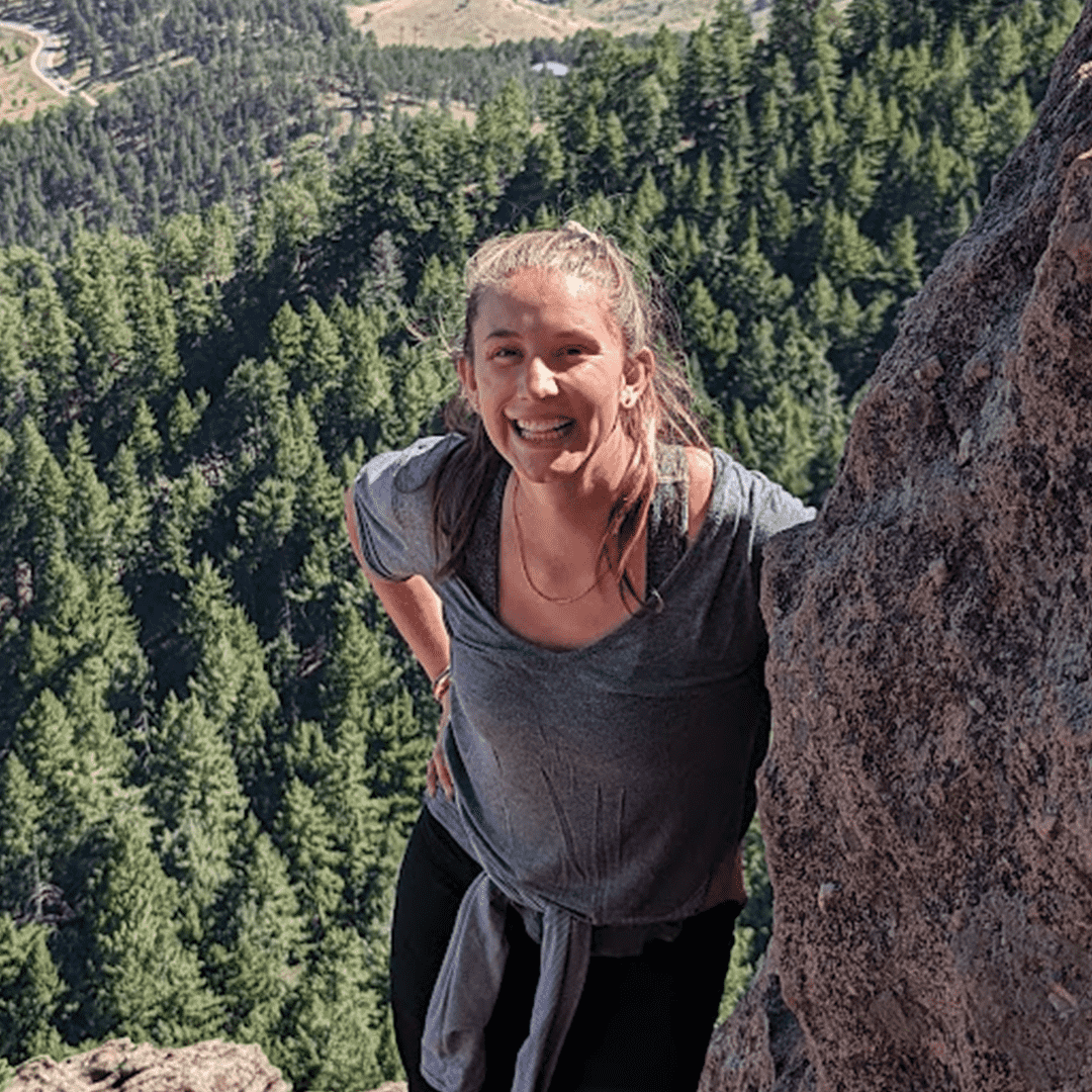 A woman smiling and leaning on a rock on a mountain with a forested landscape in the background.
