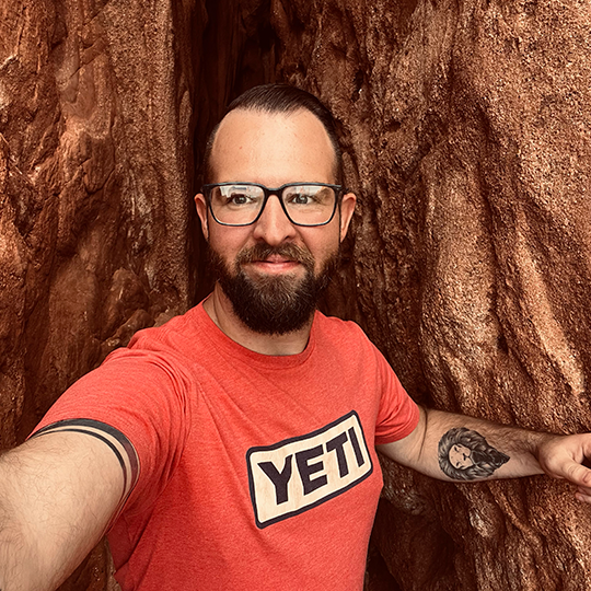 A man with glasses and a beard taking a selfie between large reddish-brown rocks.
