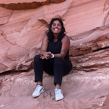 A young woman with curly hair, wearing a black sleeveless top, black pants, and white sneakers, sitting on a reddish rock formation outdoors, smiling at the camera.