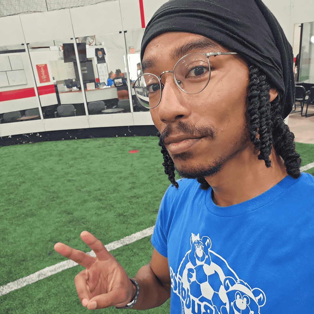 A young man with glasses and dreadlocks taking a selfie in an indoor sports facility with green artificial turf, white lines, and a glass wall with seating and people in the background.