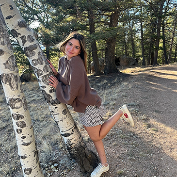 Young woman outdoors leaning against a birch tree, smiling, wearing a brown sweater, patterned shorts, and sneakers.