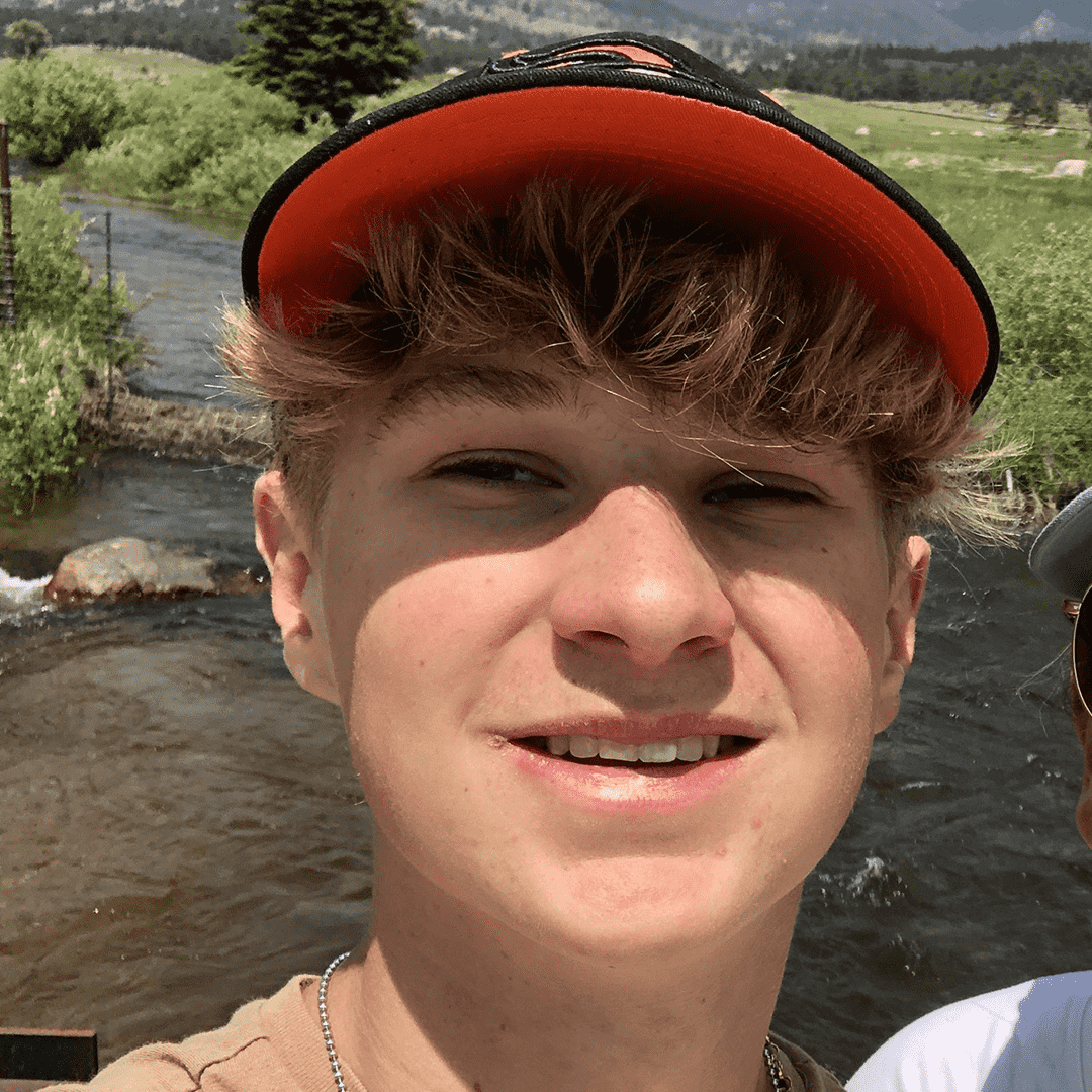 A young man with curly brown hair and a slight smile, wearing an orange and black cap, standing outdoors near a stream with green fields and hills in the background.