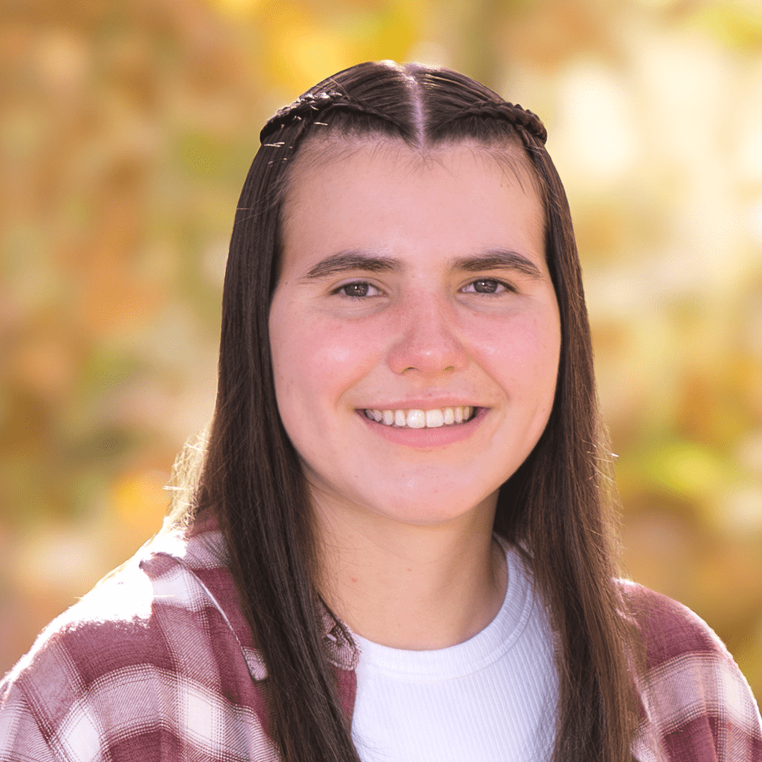 A young woman with long brown hair styled with small braids on top, smiling outdoors with a blurred background of autumn leaves.