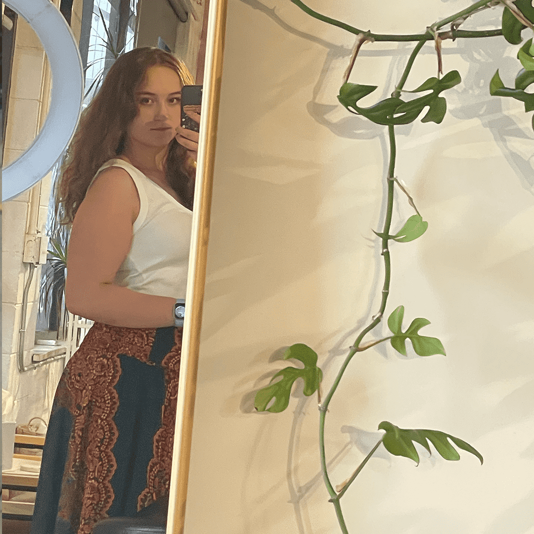 Woman taking a mirror selfie, wearing a white sleeveless top and patterned pants, with a plant and ring light visible in the background.