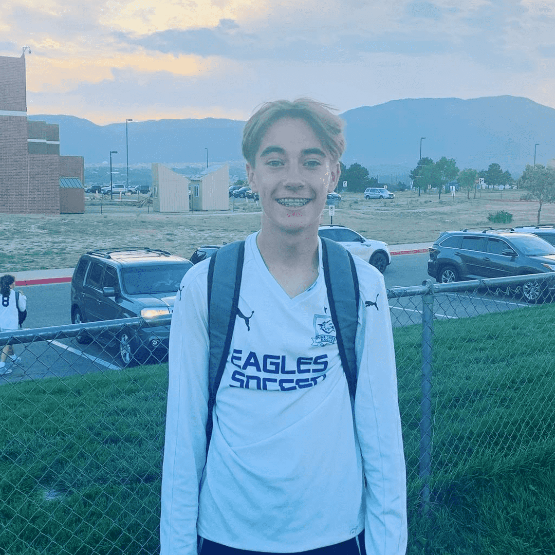 Young male soccer player smiling outdoors, wearing a white soccer jersey and backpack, standing behind a chain-link fence with parked cars and mountains in the background.