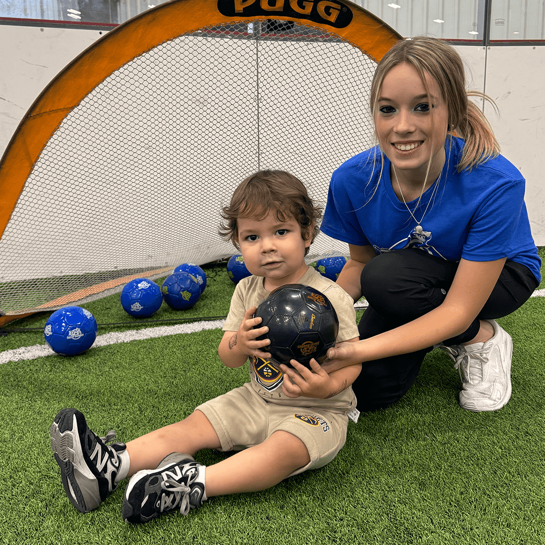 A young boy sitting on artificial turf holding a black handball, with a smiling young woman kneeling beside him, inside an indoor sports facility with a small hockey goal in the background