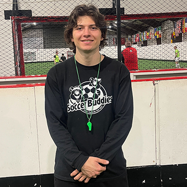 Young woman with short curly hair smiling, wearing a black 'Soccer Buddies' sweatshirt with a soccer ball graphic, standing in front of an indoor soccer field.
