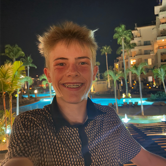 Young man with braces smiling at night near a pool with palm trees and a building in the background.